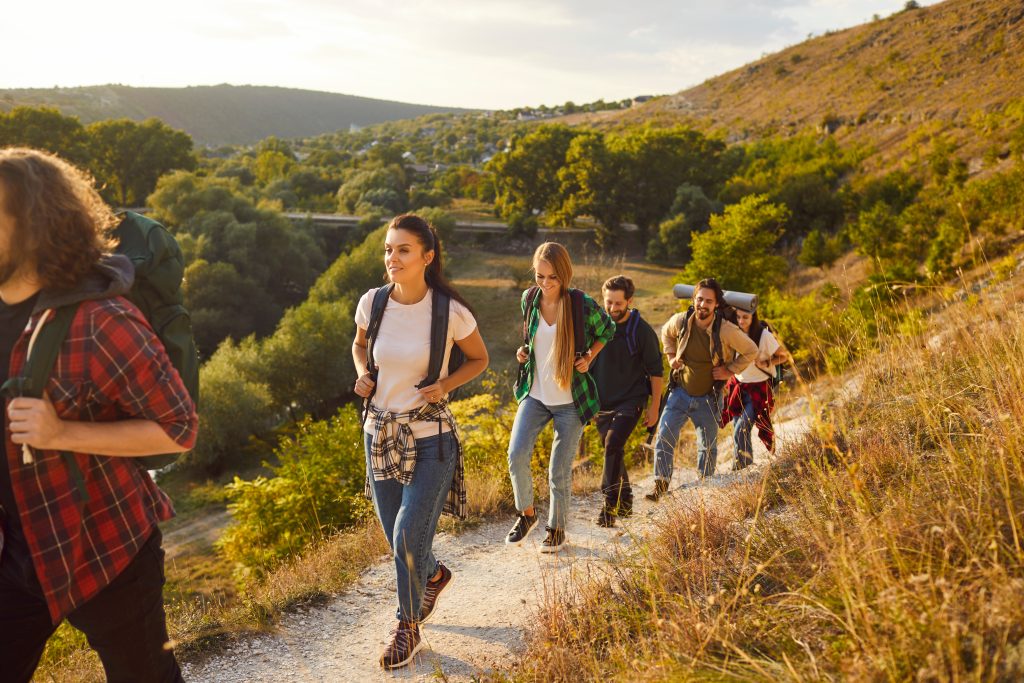 Active wanderlust people going hiking or trekking on good sunny day. Group of male female friends with backpacks following mountain trail route, leaving countryside with green woods and valleys behind